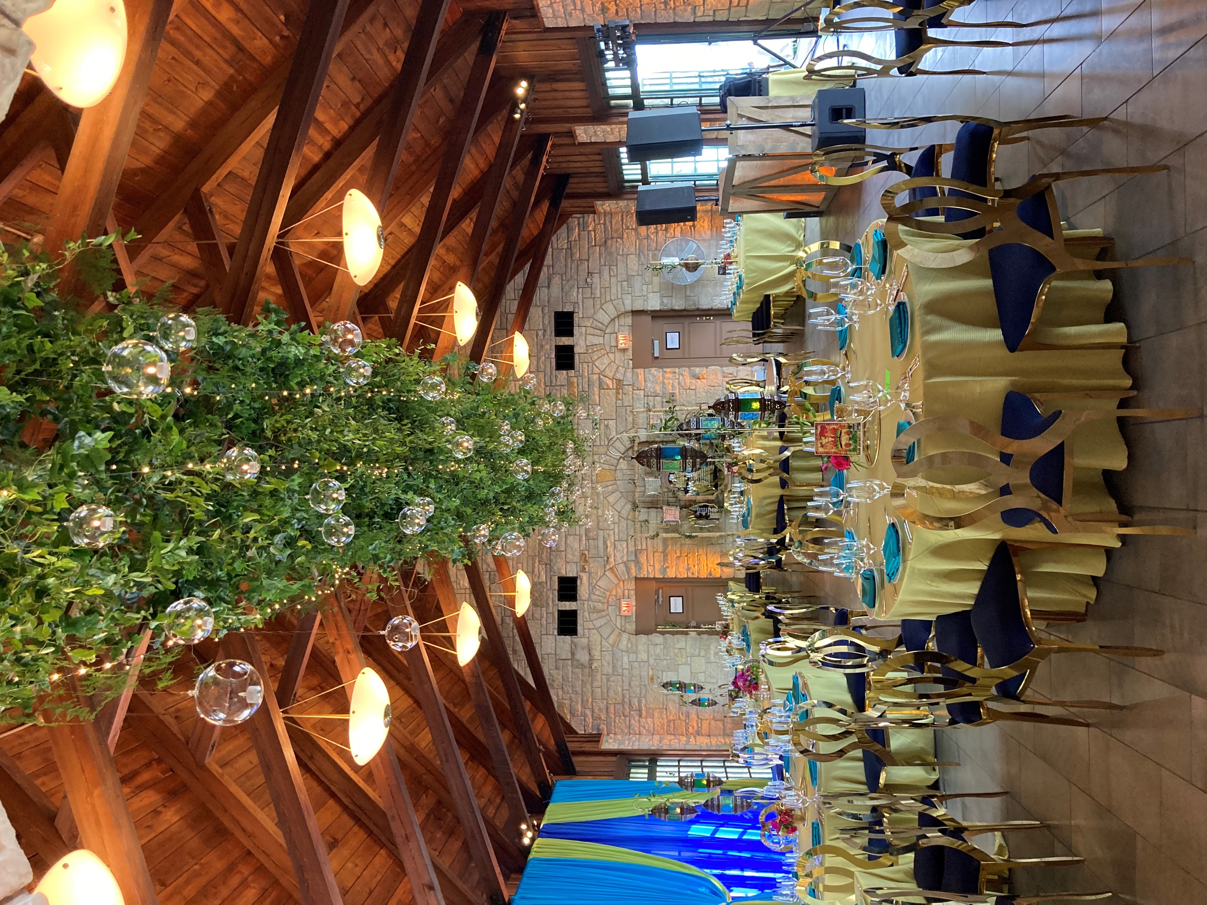 Wedding reception hall decorated with greenery, string lights, and hanging glass globes. Gold Chiavari chairs and teal and gold tablecloths add to the elegant decor.